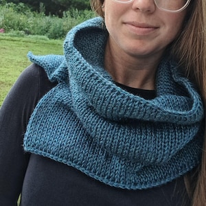 Könnte beinhalten: Ein tealblauer Strickschal. Der Schal ist aus dickem Garn gefertigt und hat ein strukturiertes Aussehen. Der Schal wird um Hals und Schultern getragen. Der Hintergrund ist eine grüne, natürliche Umgebung im Freien.