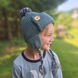 Pode incluir: Um gorro de malha verde-azulado com protetores de orelhas, um botão de madeira e um pompom. O chapéu tem duas tranças trançadas com fios azuis e bege. A pessoa está vestindo uma camisa azul clara de manga comprida com um gráfico branco.
