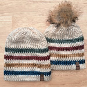 Könnte beinhalten: Zwei gestrickte Mützen mit horizontalen Streifen in Grün, Burgunderrot, Beige und Blau. Eine Mütze hat einen Kunstfell-Bommel oben. Beide Mützen sind cremefarben mit einem kleinen Lederetikett mit der Aufschrift "PVK".