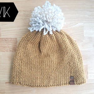 May include: Mustard yellow knit beanie with a large, fluffy, white pom-pom. The hat has a small, rectangular leather tag with the letters "PVK" on it. The beanie is made of a soft, textured yarn and is suitable for cold weather.