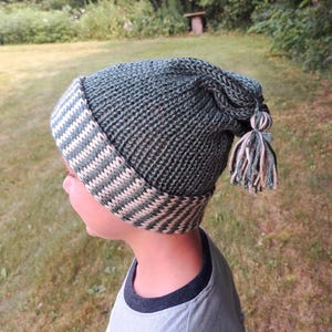 May include: A knitted green hat with a striped brim in cream and green. The hat has a tassel on the side. The hat is made of a soft, textured yarn. The hat is designed to be worn in cold weather.