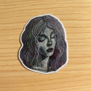 May include: A sticker featuring a grayscale illustration of a woman with closed eyes and long, wavy hair. The hair has a shimmering, iridescent effect. The woman's face is detailed with shading, and the sticker has a white border.