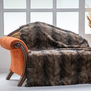 Op de afbeelding: Een bruine faux fur sprei over een oranje gestoffeerde bank met houten armen. Een kleine bijzettafel met een gouden voet en een vintage telefoon staan links. Pampasgras staat in een rieten mand.