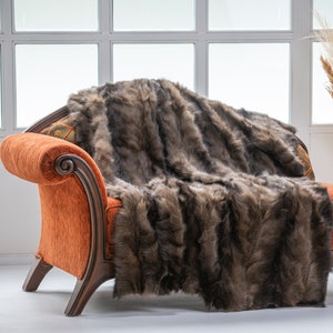 Op de afbeelding: Een bruine nep bont deken gedrapeerd over een oranje fluwelen fauteuil met houten poten. De fauteuil staat in een kamer met een witte muur en een raam erachter.