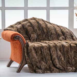 Op de afbeelding: Een bruine nep bont deken gedrapeerd over een oranje fauteuil met houten poten. De fauteuil staat in een kamer met een witte muur en een raam.