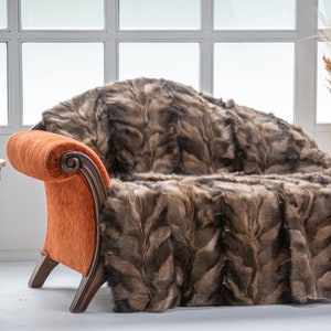 Op de afbeelding: Een bruin en grijs nep bont deken gedrapeerd over een oranje chaise longue met houten poten. De chaise longue staat in een kamer met een wit raam en een gouden bijzettafel met een vintage draaitelefoon erop.