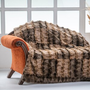 Op de afbeelding: Een bruin en beige nep bont deken gedrapeerd over een chaise longue met oranje stoffen bekleding. De chaise longue staat in een kamer met een raam en een bijzettafel met een vintage draaitelefoon.