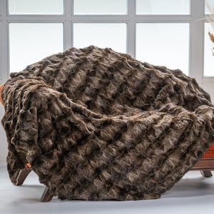 Op de afbeelding: Een bruine nep bont deken gedrapeerd over een stoel met een oranje kussen. De deken heeft een zachte, pluche textuur en is perfect om elke kamer warmte en comfort te geven.