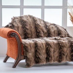 Op de afbeelding: Een bruine nep bont deken gedrapeerd over een oranje chaise longue met houten poten. De chaise longue staat in een kamer met een raam en een bijzettafel met een vintage draaitelefoon.