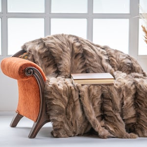 Op de afbeelding: Een bruine nep bont deken gedrapeerd over een oranje fauteuil met een boek erop. De fauteuil heeft een houten frame en een gebogen rugleuning.