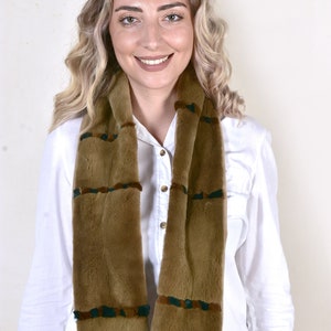 May include: A brown faux fur scarf with green and brown accents. The scarf is worn by a person with long blonde hair and a white shirt.