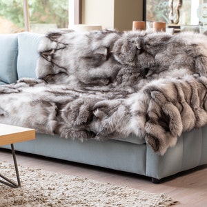 Op de afbeelding: Een luxe zilvervos bontdeken gedrapeerd over een lichtblauwe bank. De deken is zacht en pluche, met een natuurlijke, wilde uitstraling.