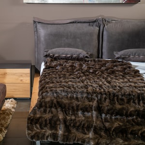 Op de afbeelding: Een moderne slaapkamer met een bed met een groot, donkergrijs hoofdbord en twee bijpassende kussens. Een zachte, bruine imitatiebont deken bedekt het bed. Een houten nachtkastje en een bijpassende poef zijn zichtbaar.