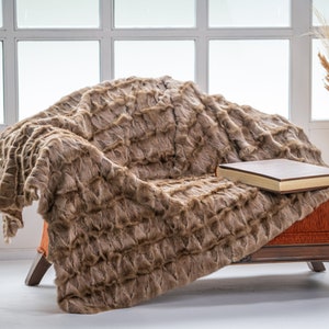 Op de afbeelding: Een bruine nep bont deken gedrapeerd over een houten fauteuil met een gesloten boek erop. De fauteuil heeft een oranje stoffen kussen en houten poten.