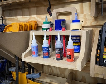 May include: A wooden wall-mounted organizer with a handle, holding bottles of glue in various colors and sizes. The organizer has a shelf for storing the bottles and a space for brushes and other tools.