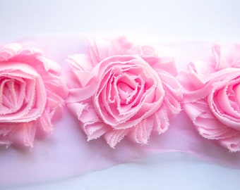 May include: Pink fabric flower trim with three large, ruffled fabric flowers. The flowers are made of soft, pink fabric and have a delicate, feminine look.