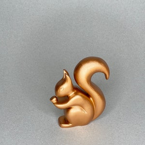 Könnte beinhalten: Eine bronzefarbene Eichhörnchenfigur, die eine Nuss hält. Die Skulptur hat eine glatte Oberfläche und ein stilisiertes Design. Der Schwanz des Eichhörnchens ist nach oben gebogen und es sitzt auf einem flachen Sockel. Die Figur ist ein dekorativer Gegenstand.