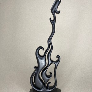 May include: A black, stylized guitar sculpture with a flame-like design. The guitar is mounted on a black, square base. The sculpture has a smooth, matte finish and is set against a neutral background.