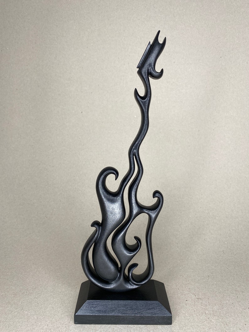May include: A black, stylized guitar sculpture with a flame-like design. The guitar is mounted on a black, square base. The sculpture has a smooth, matte finish and is set against a neutral background.