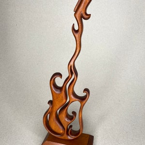 May include: A wooden sculpture of a stylized guitar in a flame-like design. The sculpture is made of a warm brown wood and stands on a rectangular base. The guitar's body is open, with flowing curves.