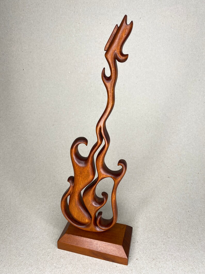 May include: A wooden sculpture of a stylized guitar in a flame-like design. The sculpture is made of a warm brown wood and stands on a rectangular base. The guitar's body is open, with flowing curves.