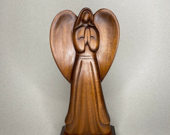 May include: A wooden angel figurine with a heart-shaped halo and hands clasped in prayer. The angel is standing on a wooden base.
