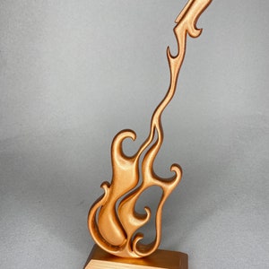 May include: A bronze-colored guitar sculpture with a flame-like design. The guitar is mounted on a rectangular base. The sculpture has a smooth, metallic finish and is designed with flowing, stylized curves.