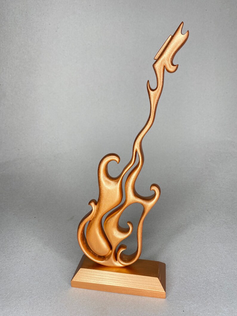 May include: A bronze-colored guitar sculpture with a flame-like design. The guitar is mounted on a rectangular base. The sculpture has a smooth, metallic finish and is designed with flowing, stylized curves.