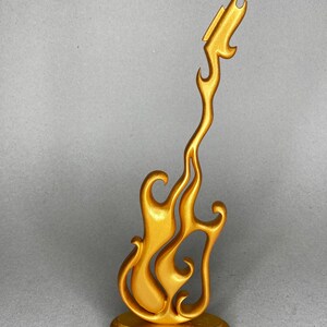 May include: A gold-colored decorative guitar sculpture with a flame-like design. The sculpture is mounted on a round base and has a smooth, metallic finish. The guitar's body is stylized with flowing curves.