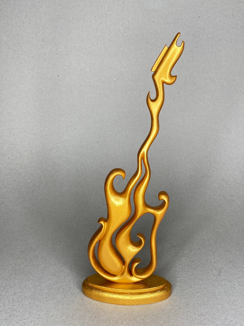 May include: A gold-colored decorative guitar sculpture with a flame-like design. The sculpture is mounted on a round base and has a smooth, metallic finish. The guitar's body is stylized with flowing curves.