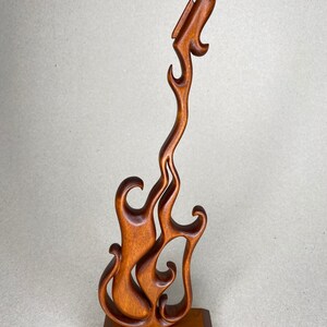 May include: A wooden sculpture of a guitar in a flame-like design. The brown sculpture is mounted on a square base. The guitar's body and neck are stylized with flowing curves, creating a dynamic and artistic representation.