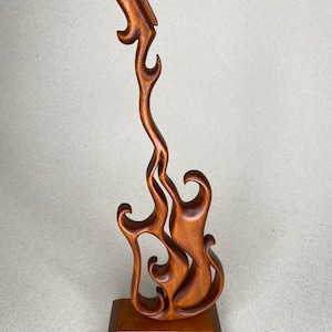 May include: A handcrafted wooden sculpture of a stylized guitar, featuring flowing, flame-like curves. The sculpture is a warm brown color and stands on a rectangular base. The design is abstract and artistic, suitable for home decor.