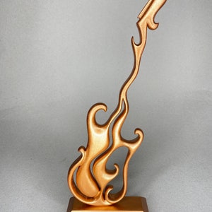 May include: A bronze-colored guitar sculpture with a flame-like design. The guitar is mounted on a rectangular base. The sculpture has a smooth, metallic finish and is set against a neutral background.