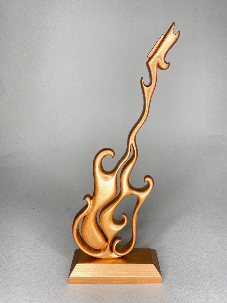 May include: A bronze-colored guitar sculpture with a flame-like design. The guitar is mounted on a rectangular base. The sculpture has a smooth, metallic finish and is set against a neutral background.