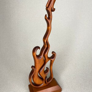 May include: A wooden sculpture of a guitar in a flame design. The brown guitar sculpture is mounted on a rectangular base. The guitar's body and neck are carved to resemble flames, creating a unique and artistic piece.