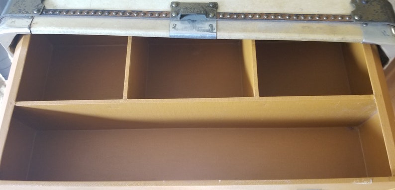 May include: The inside of a vintage suitcase with a brown fabric lining and three compartments. The compartments are separated by dividers.