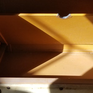 May include: The inside of a vintage white metal suitcase with a brown interior. The suitcase has a metal latch and a brown fabric lining.