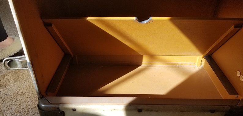 May include: The inside of a vintage white metal suitcase with a brown interior. The suitcase has a metal latch and a brown fabric lining.