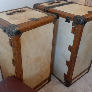 May include: Two vintage suitcases with brown leather trim and brass hardware. The suitcases are made of wood and have a light beige finish.