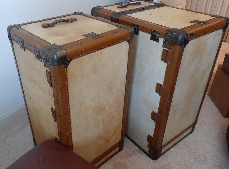 May include: Two vintage suitcases with brown leather trim and brass hardware. The suitcases are made of wood and have a light beige finish.
