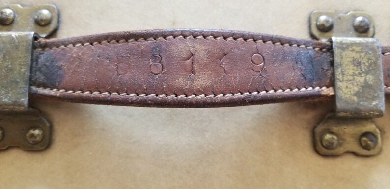 May include: Close-up of a brown leather handle with the numbers "319" embossed on it. The handle is attached to a vintage suitcase.