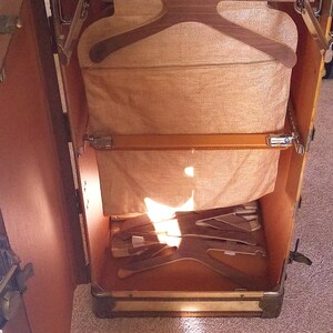 May include: An open vintage wooden suitcase with a brown leather exterior and a beige fabric interior. The suitcase has a built-in hanger system with wooden hangers and a fabric lining.