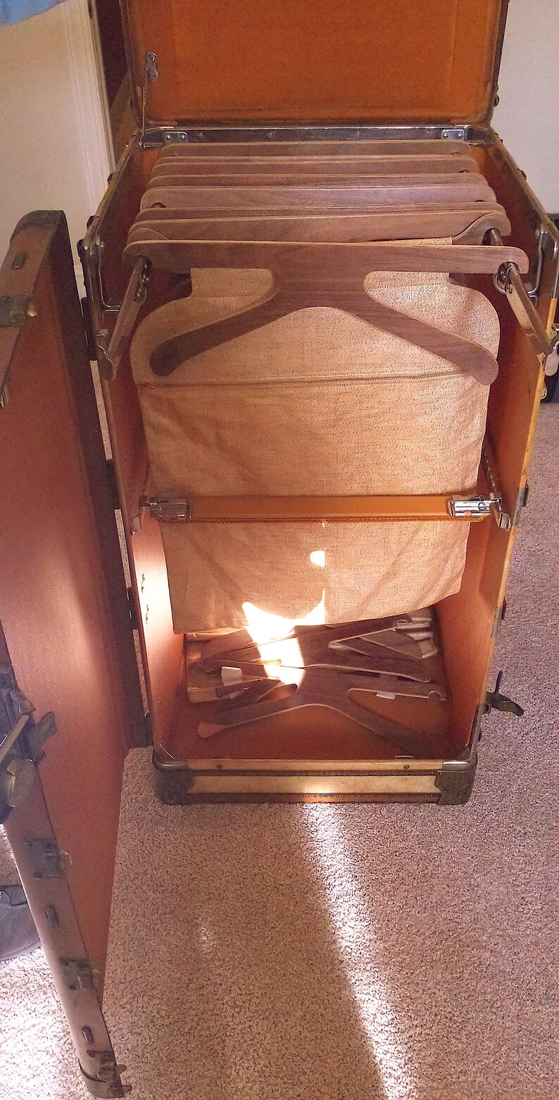 May include: An open vintage wooden suitcase with a brown leather exterior and a beige fabric interior. The suitcase has a built-in hanger system with wooden hangers and a fabric lining.