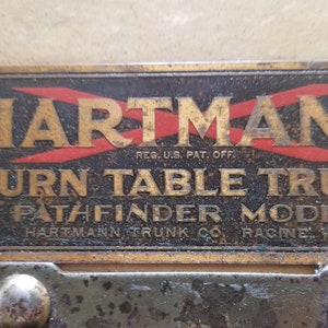 May include: A brass plaque with the text "HARTMANN" in gold lettering with red diagonal lines, "REG. U.S. PAT. OFF." in smaller lettering, "TURN TABLE TRUNK" in gold lettering, "PATHFINDER MODEL" in gold lettering, and "HARTMANN TRUNK CO. RACINE WIS." in smaller lettering.