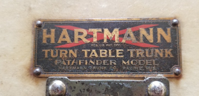 May include: A brass plaque with the text "HARTMANN" in gold lettering with red diagonal lines, "REG. U.S. PAT. OFF." in smaller lettering, "TURN TABLE TRUNK" in gold lettering, "PATHFINDER MODEL" in gold lettering, and "HARTMANN TRUNK CO. RACINE WIS." in smaller lettering.