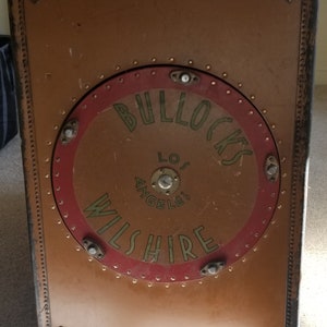 May include: A vintage brown suitcase with a circular logo that reads "Bullocks Wilshire Los Angeles" in gold lettering on a red background.