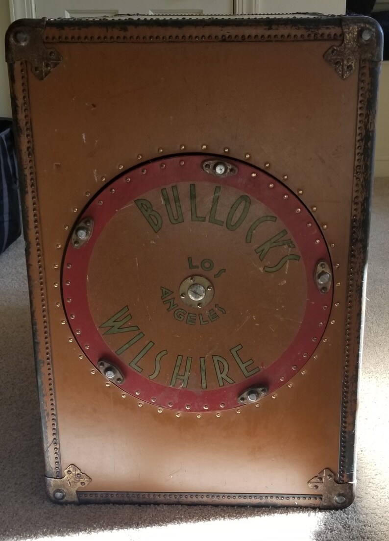 May include: A vintage brown suitcase with a circular logo that reads "Bullocks Wilshire Los Angeles" in gold lettering on a red background.
