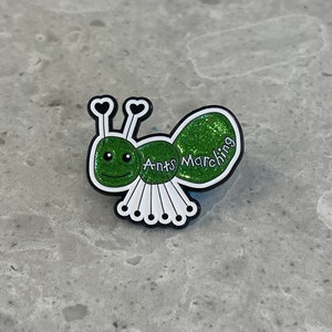 May include: A green glitter ant pin with white legs and black antennae. The ant has a smiling face and the words "Ants Marching" are written on its body.