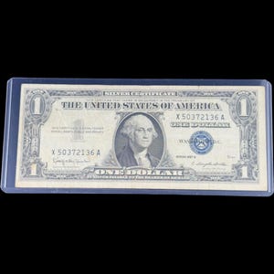 May include: A one-dollar silver certificate from the United States of America, featuring a portrait of George Washington. The bill is encased in a protective sleeve. The text "Silver Certificate" is visible at the top.