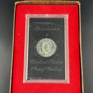 May include: A 1971 Eisenhower silver dollar coin in a protective case. The coin features a profile of President Eisenhower and the words "IN GOD WE TRUST." The coin is displayed on a black background within a red velvet-lined box.